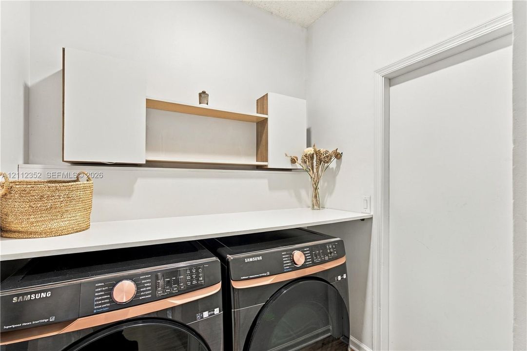 Laundry area featuring built-in white cabinetry, an open wood-finish shelf, and a white countertop above laundry appliances