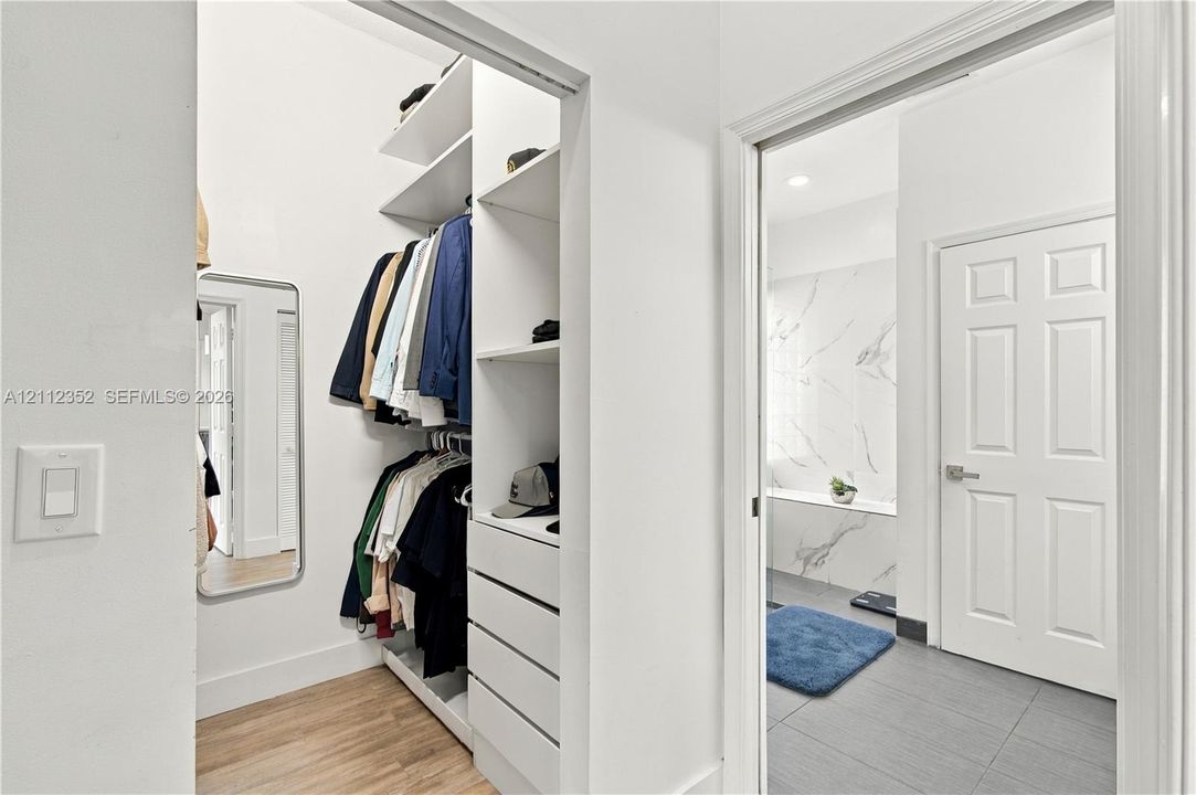 Walk-in closet featuring custom white shelving, integrated drawers, and wood-finish flooring, accompanied by a bathroom showcasing a stone-tile shower surround and large-format floor tile