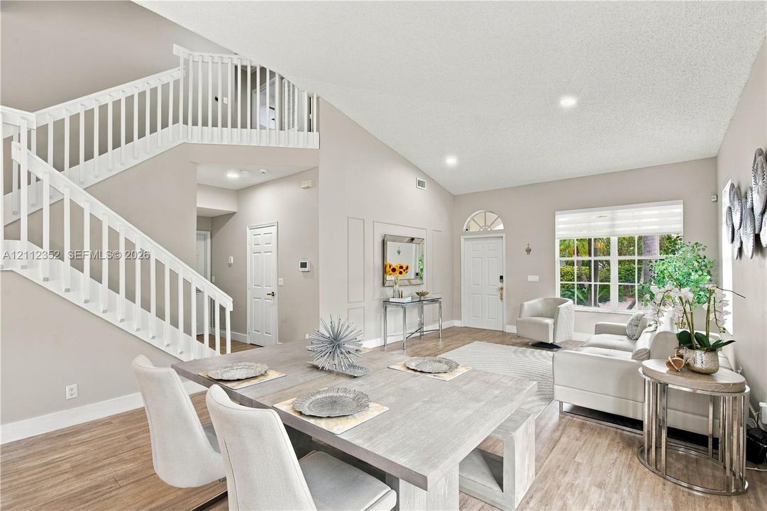 Open-concept interior featuring vaulted ceilings, wood-finish flooring, a white staircase with decorative balustrade, and multi-paned windows with a transom