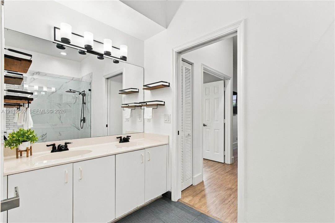 Dual-sink vanity with solid-surface countertop, full-width wall mirror, four-light vanity fixture, matte black hardware, and shower featuring large-format wall tile