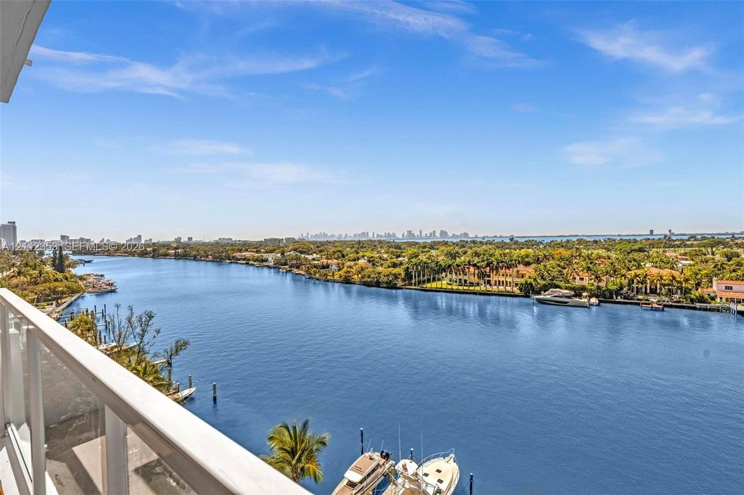 Balcony featuring a white metal railing overlooking a wide waterway, coastal vegetation, and a distant city skyline