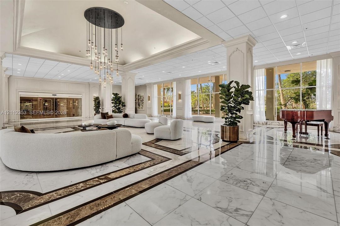 Grand lobby featuring polished stone flooring with decorative inlay, soaring tray ceilings, an intricate multi-pendant chandelier, structural pillars, and expansive floor-to-ceiling windows