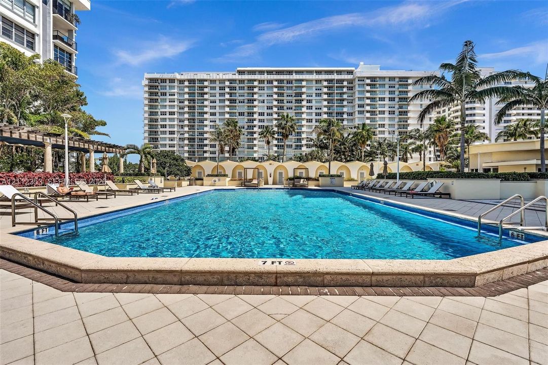 Rectangular swimming pool featuring a stone-tile deck, perimeter stone coping, metal safety rails, and integrated poolside cabanas