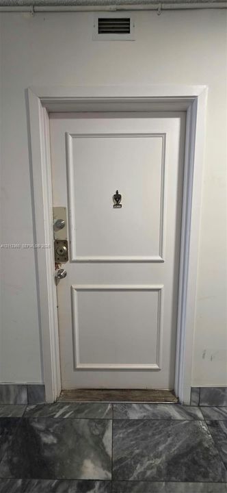 White paneled door featuring a decorative metal door knocker, dual deadbolt locks, and a polished metal door handle, set against stone-finish flooring