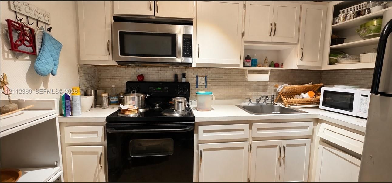 Kitchen featuring white shaker-style cabinetry, a stone tile backsplash, light-toned countertops, a stainless steel over-the-range microwave, and an integrated basin sink