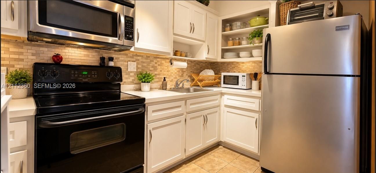 Kitchen featuring white cabinetry, stainless steel appliances, neutral-toned stone tile backsplash, integrated open shelving, and solid-surface countertops
