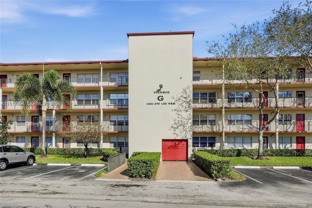 Multi-story residential building exterior featuring a central stucco facade, horizontal balconies with metal railings, red-accented doors, and asphalt parking area
