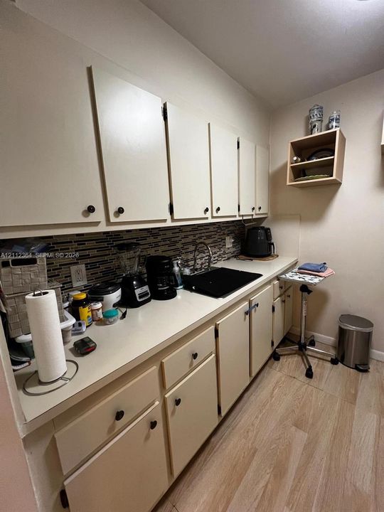 Kitchen featuring solid-color cabinetry, neutral-toned countertops, multi-toned linear tile backsplash, dark-finished integrated sink, and wood-finish flooring