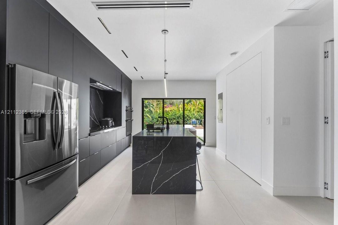 Contemporary kitchen featuring matte charcoal cabinetry, a waterfall-edge stone island, stainless steel refrigerator, built-in double ovens, and large-format tile flooring