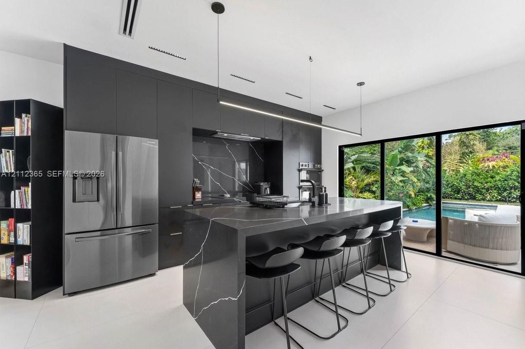 Modern kitchen featuring matte cabinetry, stone-surfaced island with waterfall edge, integrated stainless steel refrigerator, linear pendant lighting, and full-height sliding glass doors