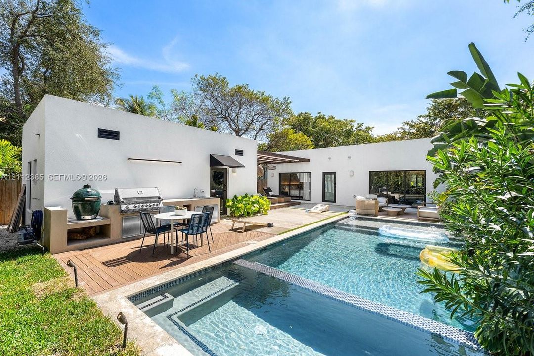 Outdoor living area featuring a built-in kitchen with stainless steel grill, composite wood decking, in-ground swimming pool with tiled border, and modern stucco exterior