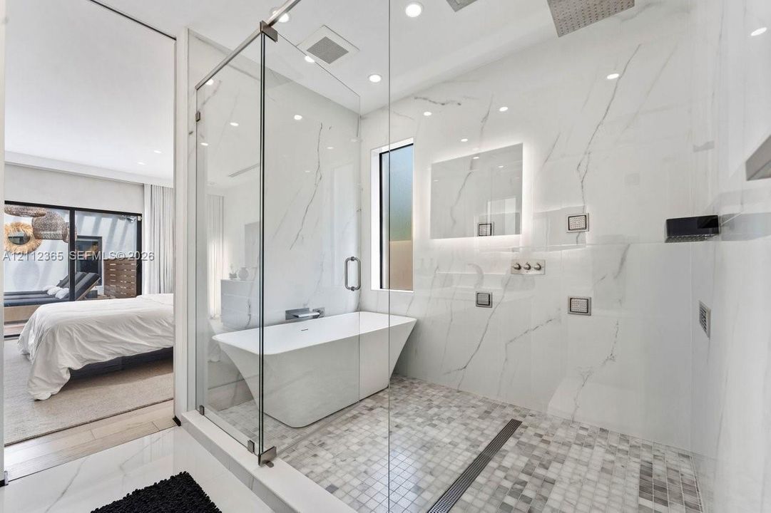 Walk-in wet room featuring a freestanding soaking tub, frameless glass enclosure, floor-to-ceiling stone-finish wall tile, mosaic tile flooring, and wall-mounted plumbing fixtures