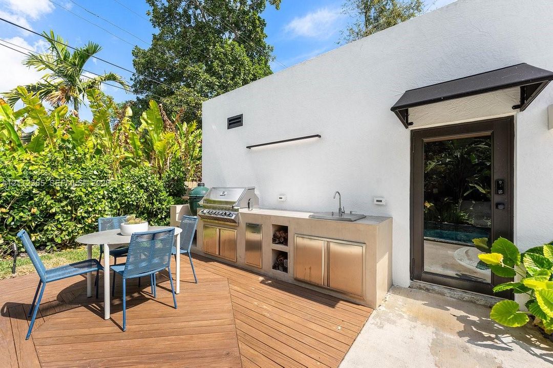 Outdoor kitchen featuring a built-in stainless steel grill, integrated sink, and cabinetry set on a wood-finish deck with a glass-paneled exterior door and black metal awning