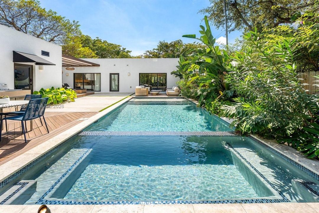 In-ground lap pool with an integrated spa, stone coping, wood-finish decking, and white stucco exterior walls