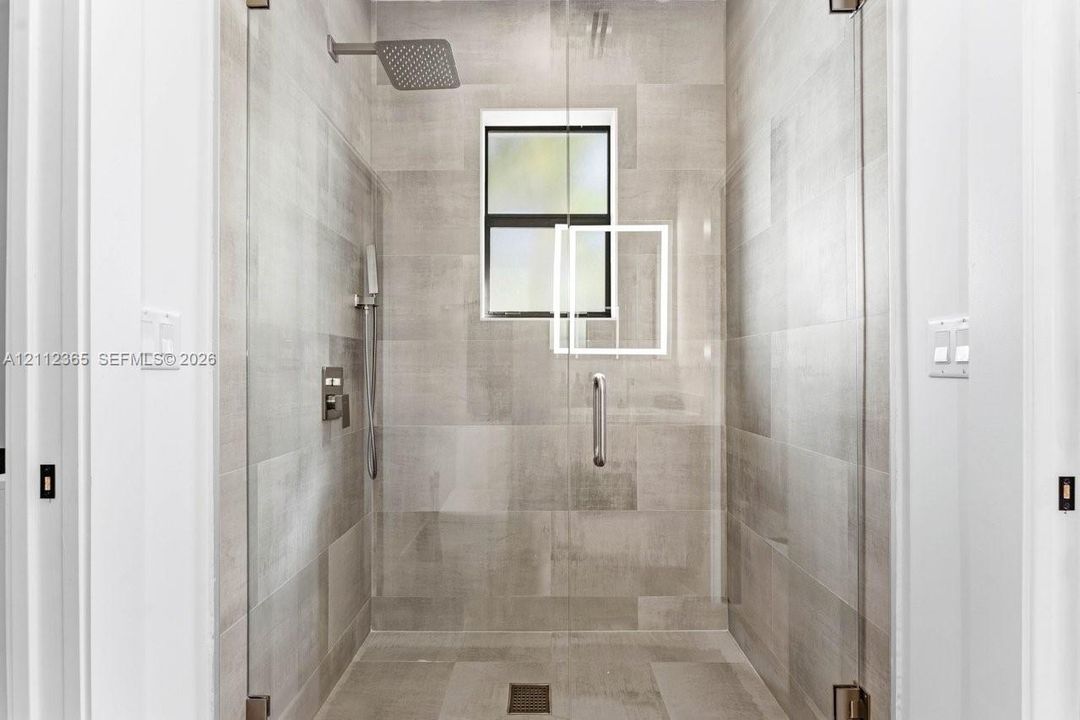 Walk-in shower featuring large-format neutral tile, frameless glass enclosure, rainfall shower head, handheld sprayer, and built-in window