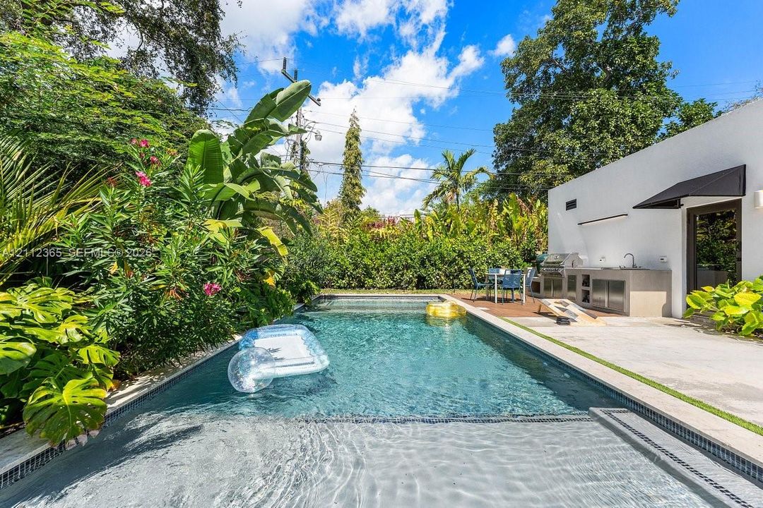 Swimming pool with a baja shelf, concrete patio, built-in outdoor kitchen with sink, and lush perimeter landscaping