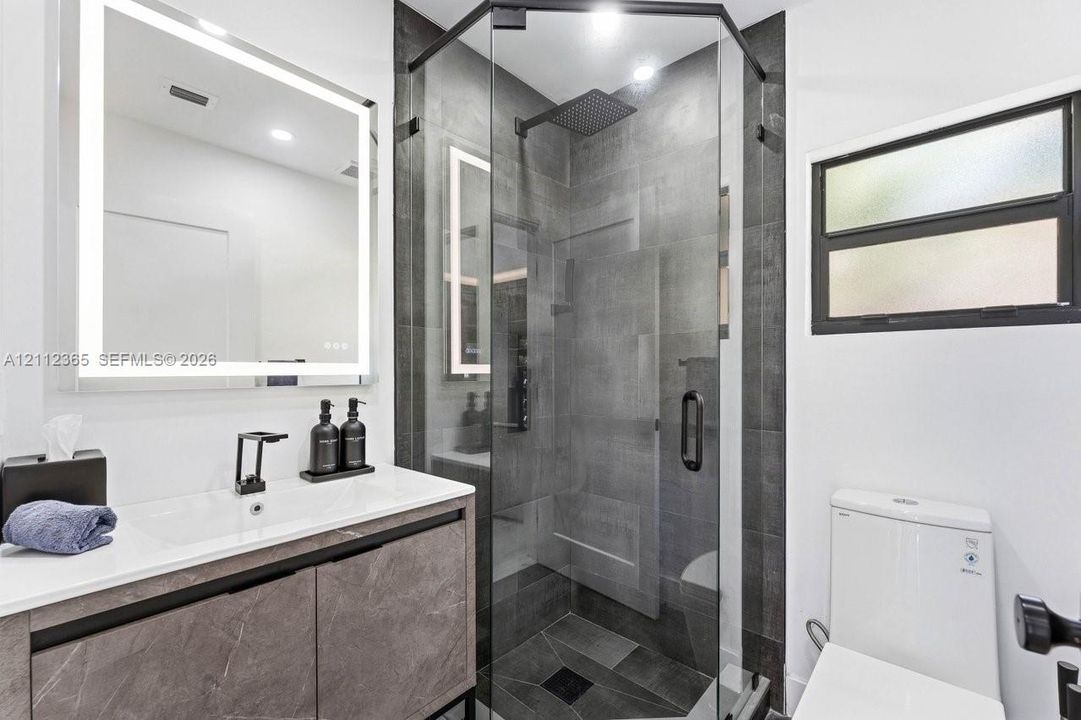 Bathroom featuring a floating vanity with a stone-finish cabinet, integrated LED-backlit mirror, glass-enclosed shower with dark tile surround, and black plumbing fixtures