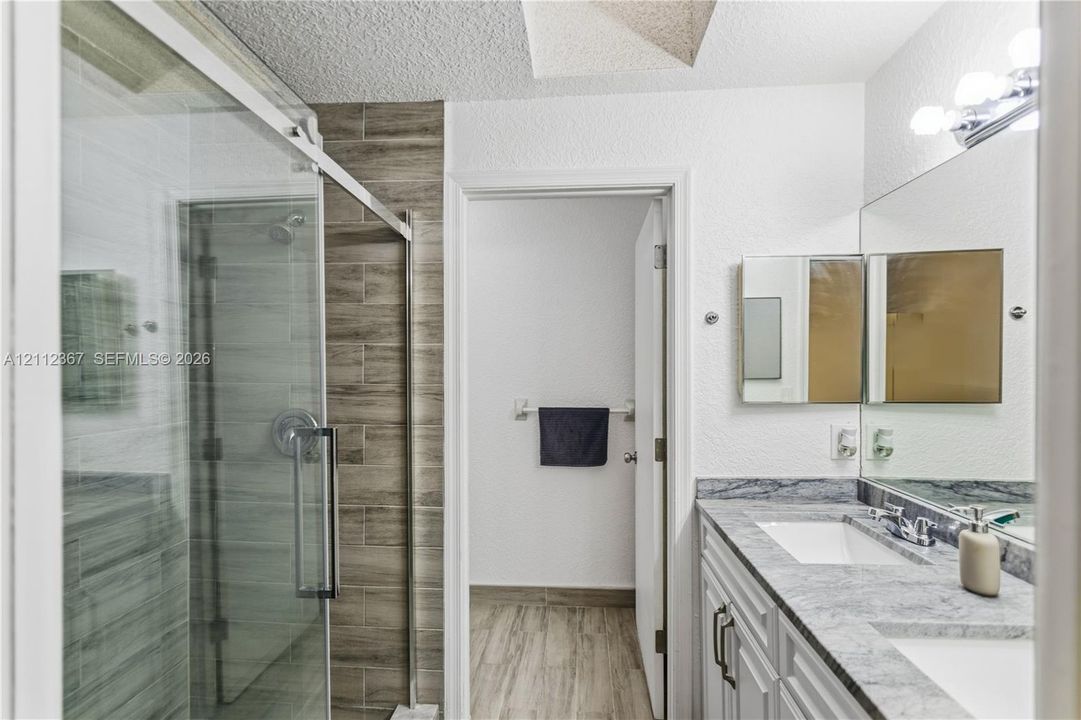 Bathroom featuring a dual-sink vanity with stone-finish countertop, walk-in shower with glass enclosure, and wood-finish tile surround