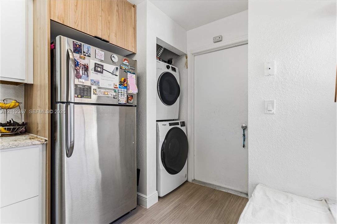 Stainless steel refrigerator with overhead custom cabinetry, stacked front-load laundry appliances, and wood-finish flooring