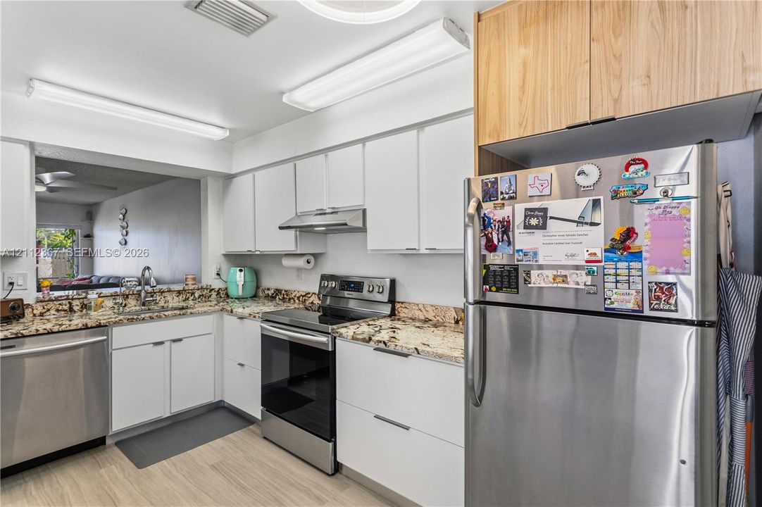Kitchen featuring white flat-panel cabinetry, light-toned wood-finish upper cabinets, stone-surfaced countertops, stainless steel appliances, and wood-finish flooring