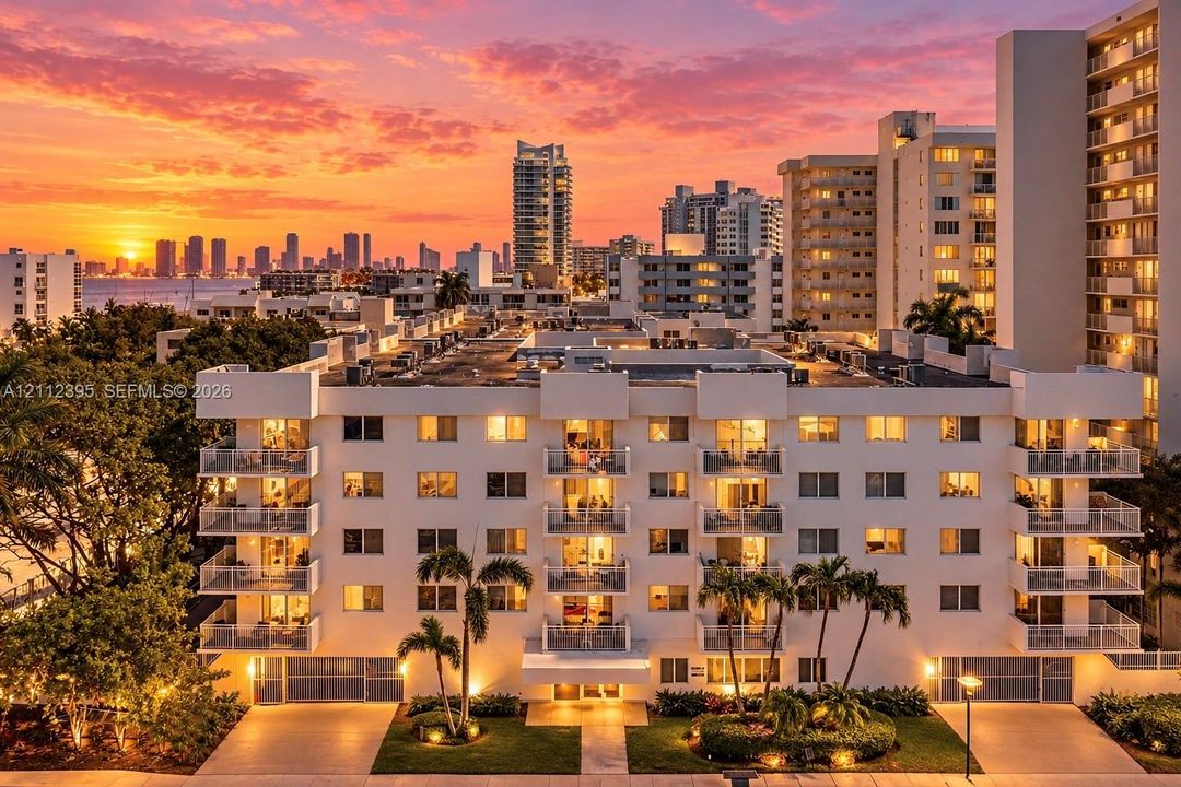 Mid-rise residential building featuring stacked private balconies with glass railings, landscaped grounds with mature palm trees, and exterior hardscaping