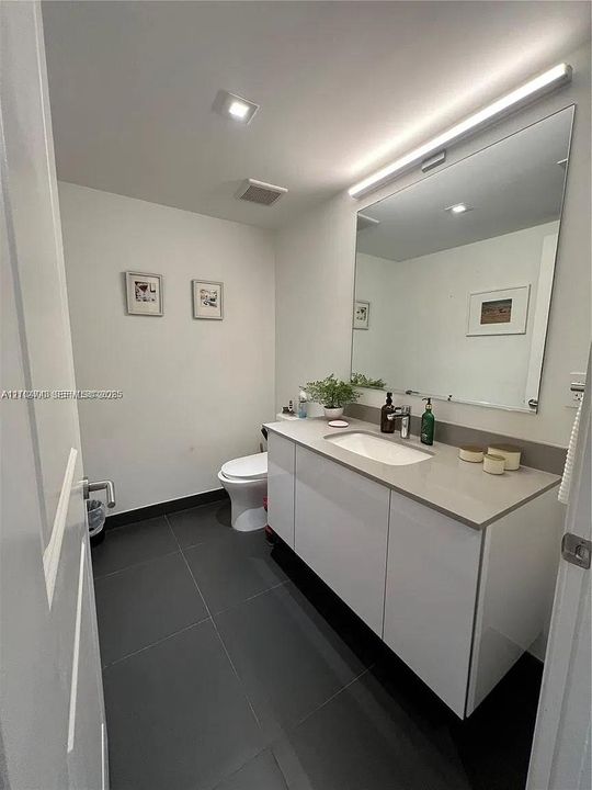 Floating white vanity with solid-surface countertop and integrated sink, large frameless vanity mirror with integrated horizontal light fixture, and dark-toned floor tiling