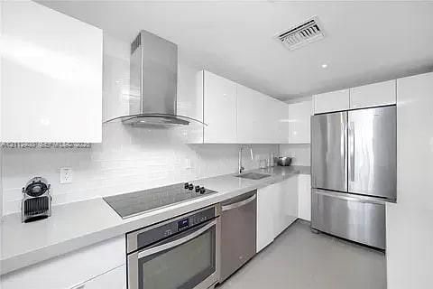 Modern kitchen featuring flat-panel high-gloss cabinetry, stone countertops, stainless steel appliances, a tile backsplash, and a wall-mounted chimney-style range hood