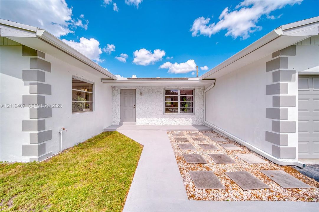 Exterior featuring white stucco finish with grey decorative quoins, textured stone masonry accents, a concrete walkway, and a geometric hardscape design