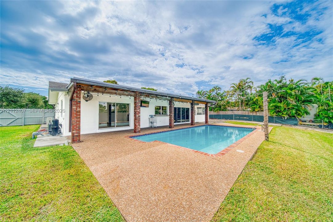 Rear exterior featuring a covered patio with brick support columns, an in-ground pool with brick coping, expansive lawn, and fenced perimeter