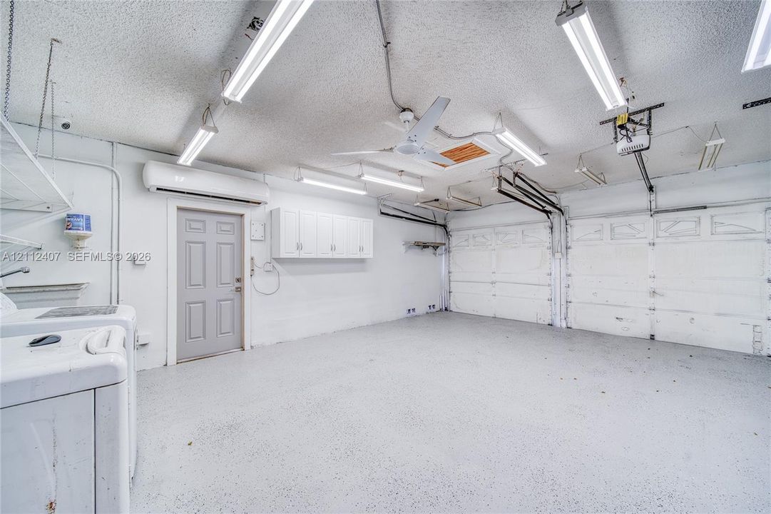 Garage interior featuring epoxy-coated flooring, wall-mounted cabinetry, a ceiling-mounted fan, overhead fluorescent lighting, and a climate-control unit