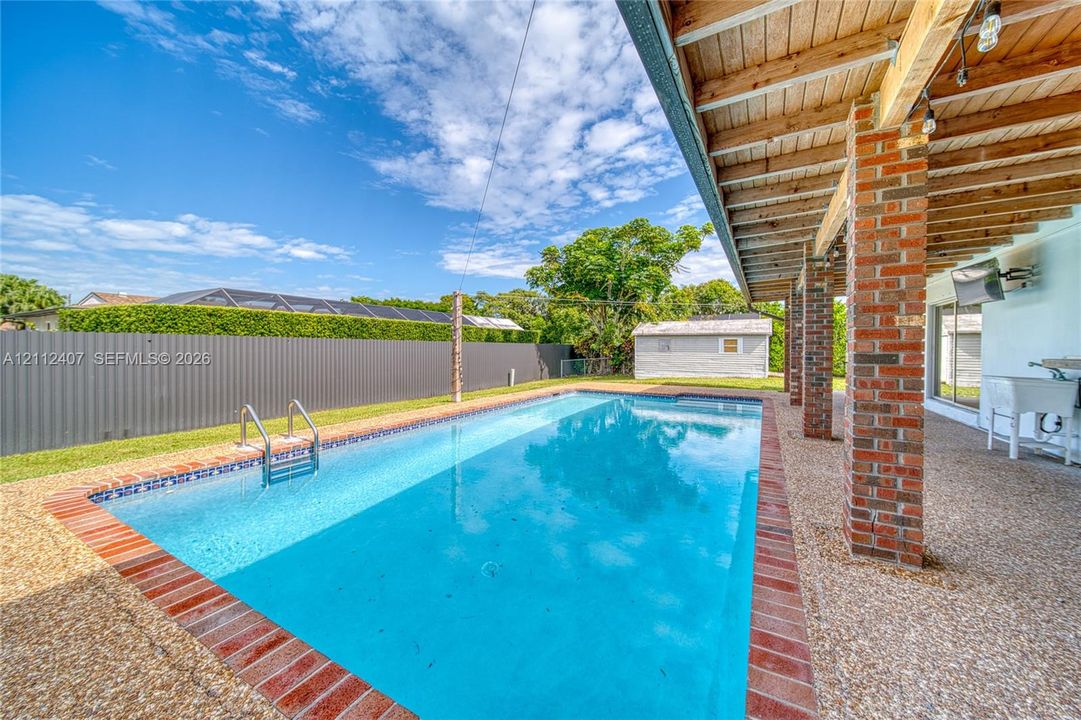 Rectangular in-ground swimming pool featuring a brick surround, covered patio with exposed wood ceiling beams, exposed brick structural pillars, and a metal privacy fence
