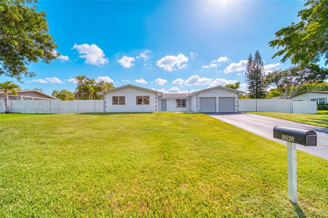 Single-story exterior featuring a painted masonry facade, two-car garage, asphalt driveway, and full perimeter vinyl fencing on a grass lot
