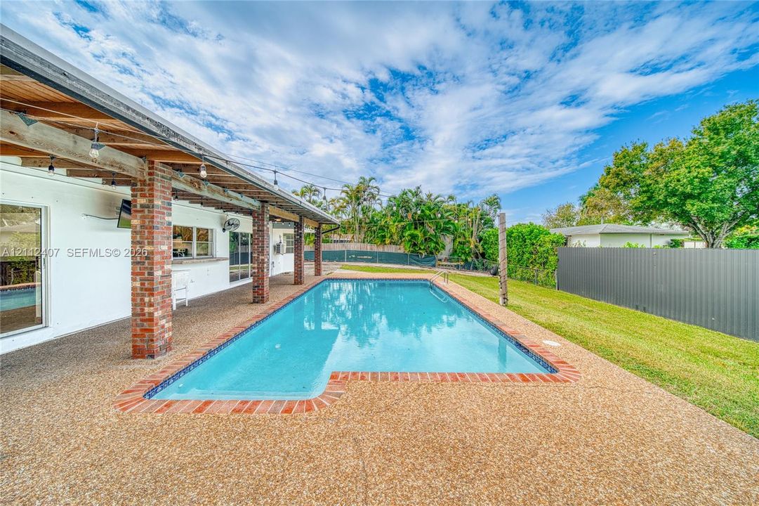 Covered outdoor patio with exposed wood beams and brick support columns, in-ground swimming pool with brick coping, and a fenced backyard