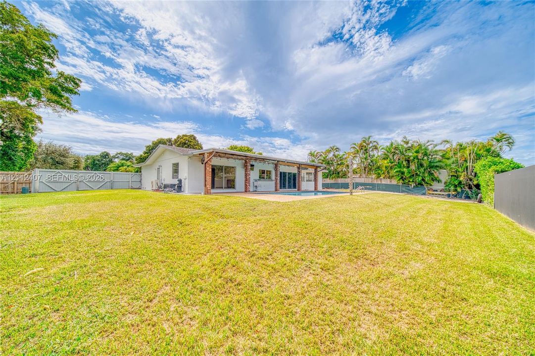 Expansive backyard featuring a large grass lawn, perimeter fencing, an in-ground pool, and a covered patio with brick structural supports