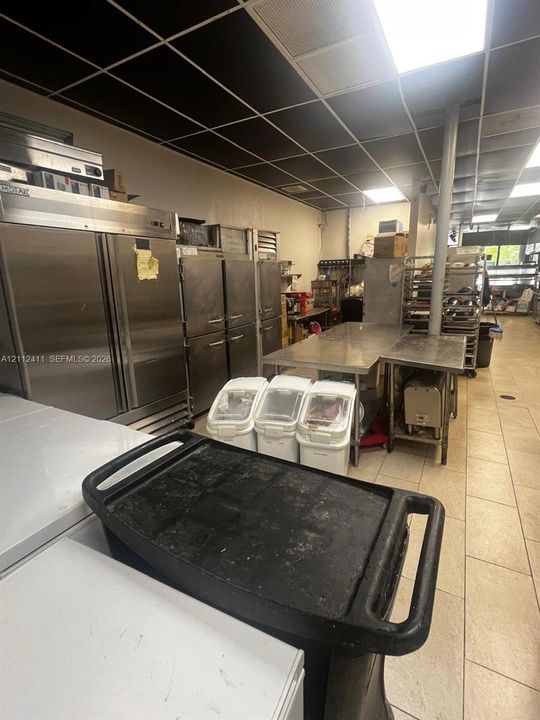 Commercial kitchen space featuring stainless steel refrigeration units, stainless steel prep tables, tile flooring, and drop ceiling panels with integrated lighting