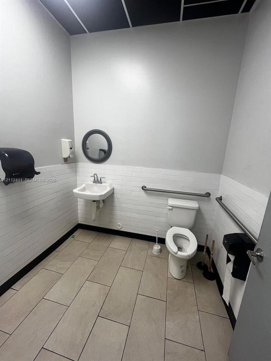 Restroom featuring large-format ceramic tile flooring, horizontal subway tile wainscoting, wall-mounted sink, and metal safety grab bars