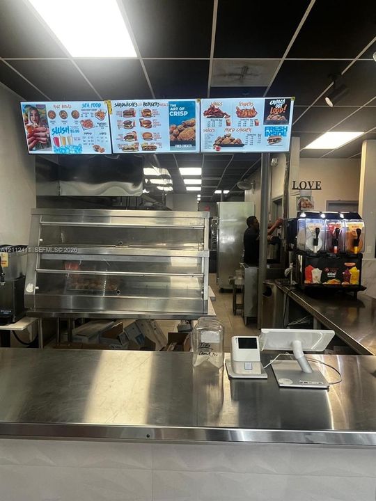 Commercial kitchen space featuring stainless steel food preparation counters, a multi-tier heated service display, suspended menu signage, and recessed ceiling lighting