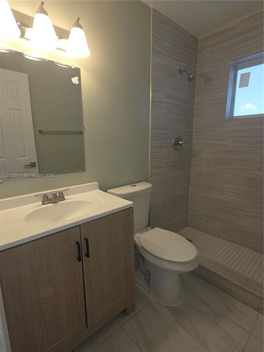 Wood-finish vanity with a composite countertop, wall-mounted mirror, light fixture, walk-in shower with horizontal tile surround, and tile flooring