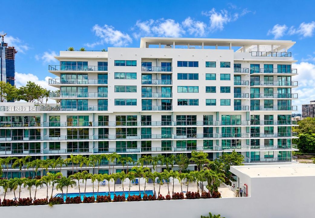 Mid-rise residential building featuring floor-to-ceiling glass windows, private balconies with glass railings, a rooftop terrace, and an outdoor swimming pool surrounded by landscaped palm trees