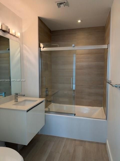 Modern floating vanity with a light-toned surface, tub-shower combination featuring a glass sliding door, wood-finish wall tile surround, and wood-finish flooring