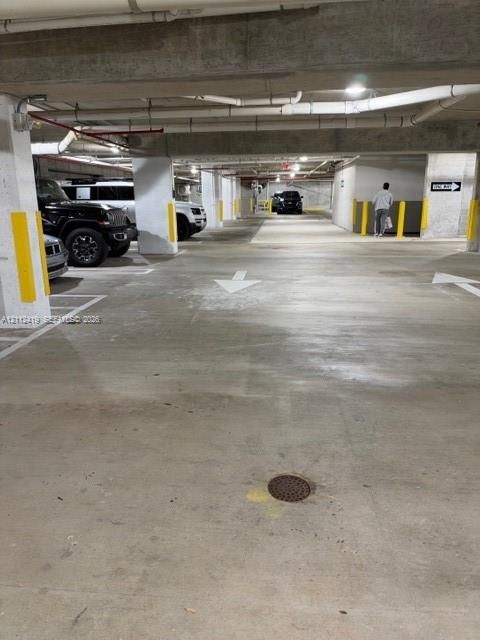 Covered parking garage with concrete flooring, structural pillars featuring yellow protective markers, and painted directional signage