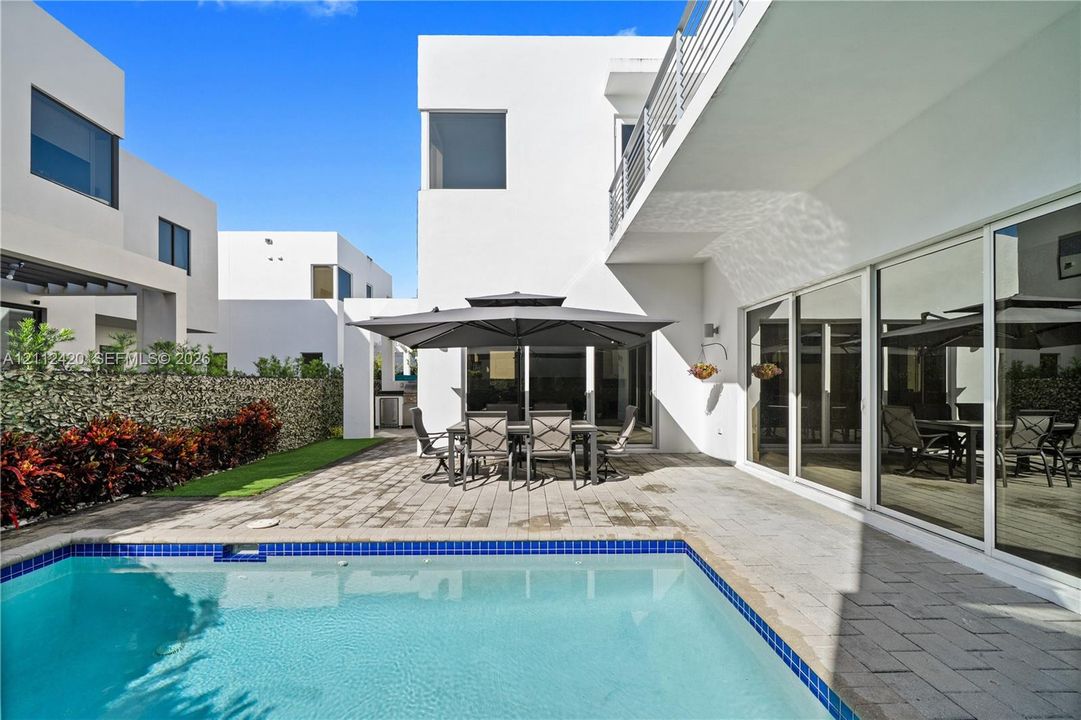 Modern stucco exterior with expansive sliding glass doors, paver patio, in-ground pool with blue tile border, and privacy hedge