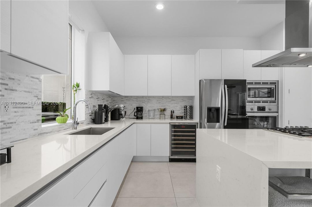 Modern kitchen featuring white flat-panel cabinetry, quartz countertops, a tiled backsplash, stainless steel appliances including a built-in wine refrigerator, and a waterfall-edge kitchen island