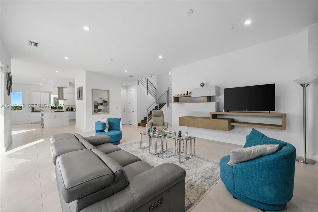 Open-concept living space featuring large-format tile flooring, recessed lighting, wall-mounted floating cabinetry, and a staircase with metal horizontal railings