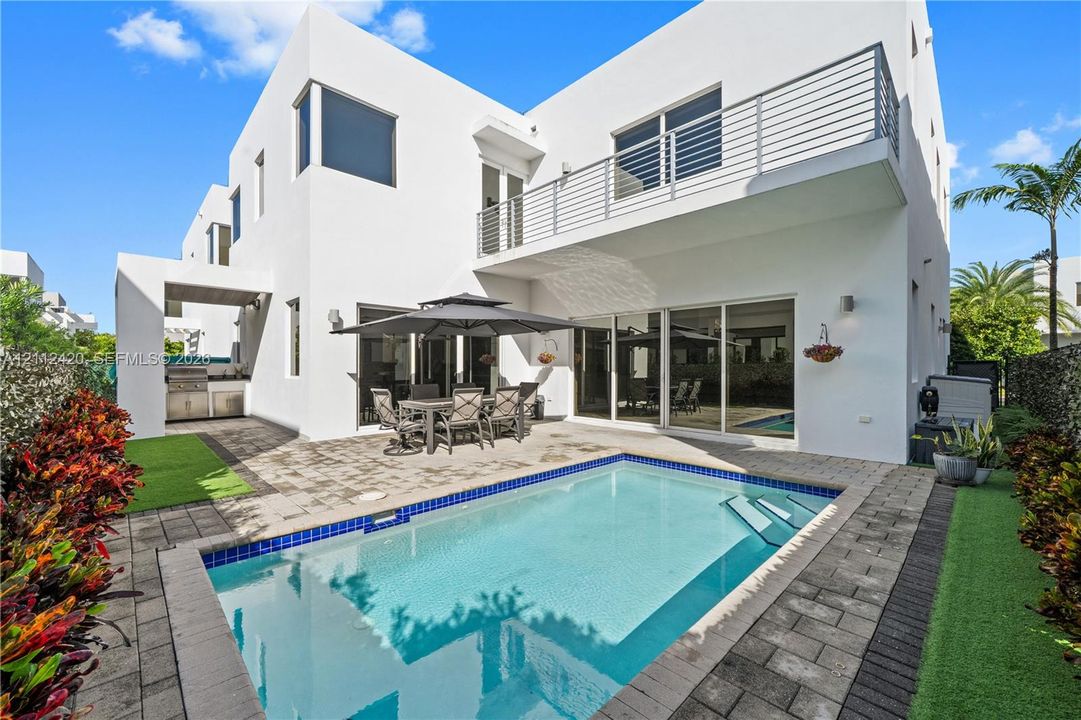 Modern two-story stucco exterior featuring a built-in outdoor kitchen, second-level balcony with horizontal metal railings, concrete paver patio, and an in-ground swimming pool