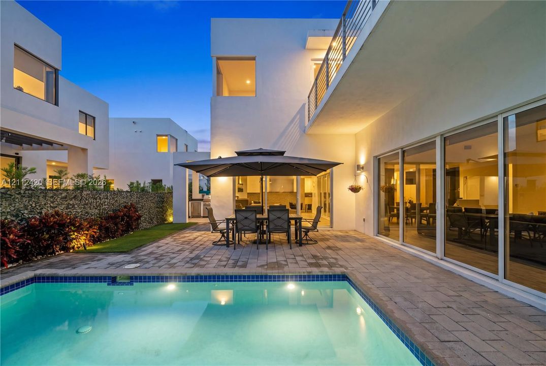 Modern residence featuring a swimming pool with tile trim, expansive stone paver patio, minimalist stucco exterior, and multi-panel floor-to-ceiling glass sliding doors