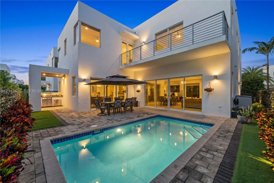 Modern two-story residence featuring a paved pool deck, custom swimming pool, exterior kitchen with stainless steel grill, and an upper-level balcony with metal railing