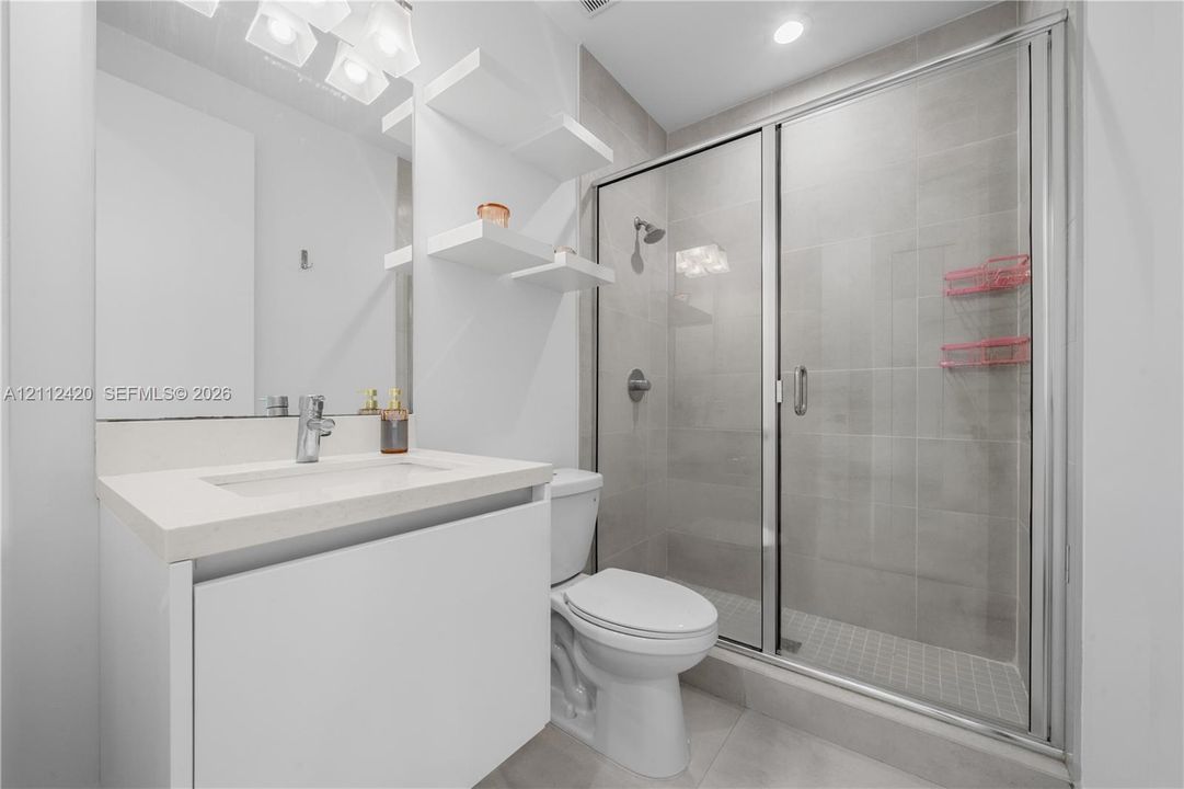 Bathroom featuring a white vanity with a rectangular sink, quartz countertop, built-in wall shelving, and a tiled shower enclosure with a glass door