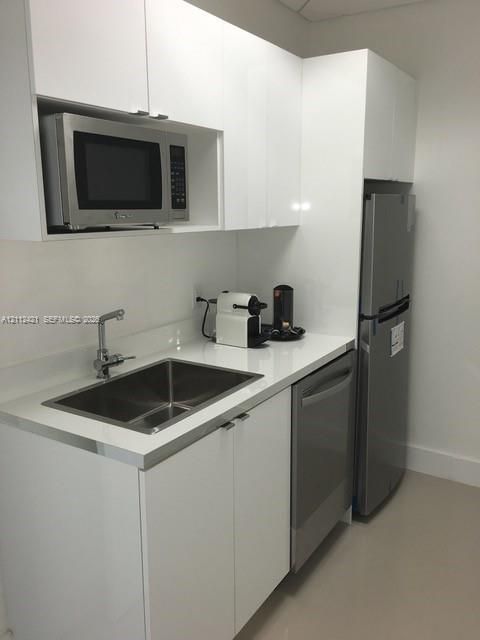 Minimalist kitchenette featuring high-gloss white cabinetry, solid-surface countertops, integrated stainless steel sink with gooseneck faucet, and stainless steel appliances