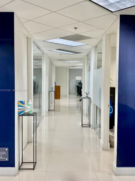 Hallway featuring high-gloss tile flooring, suspended acoustic ceiling panels, inset fluorescent lighting, and glass-partitioned wall segments